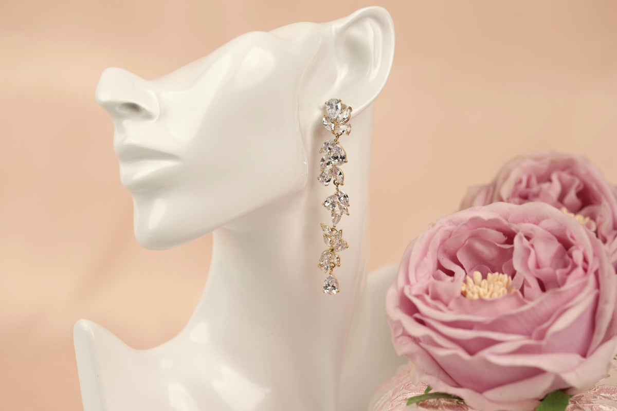 A white mannequin head wearing a pair of gold dangling earrings with clear gemstones, placed next to a bouquet of pink roses.