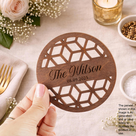 A hand holding a wooden coaster with the name 'The Wilson' and the date '08.09.2025' engraved on it, surrounded by various tableware and decorative items.