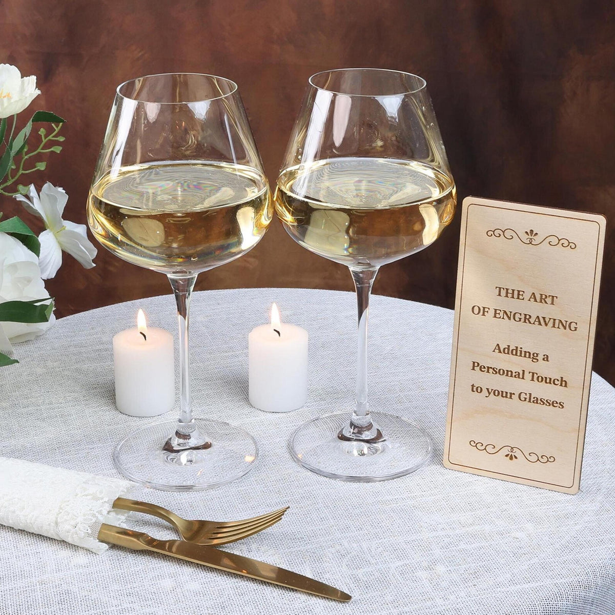 two wine glasses filled with white wine, accompanied by a wooden sign that reads "The Art of Engraving: Adding a Personal Touch to your Glasses".