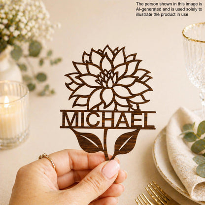 A hand holding a wooden cutout of a flower with the name "Michael" written on it, against a backdrop of floral arrangements and a glass of wine.