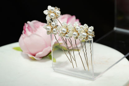 A clear display case containing several ornate hair clips with white pearls and gold accents, placed next to a pink rose.