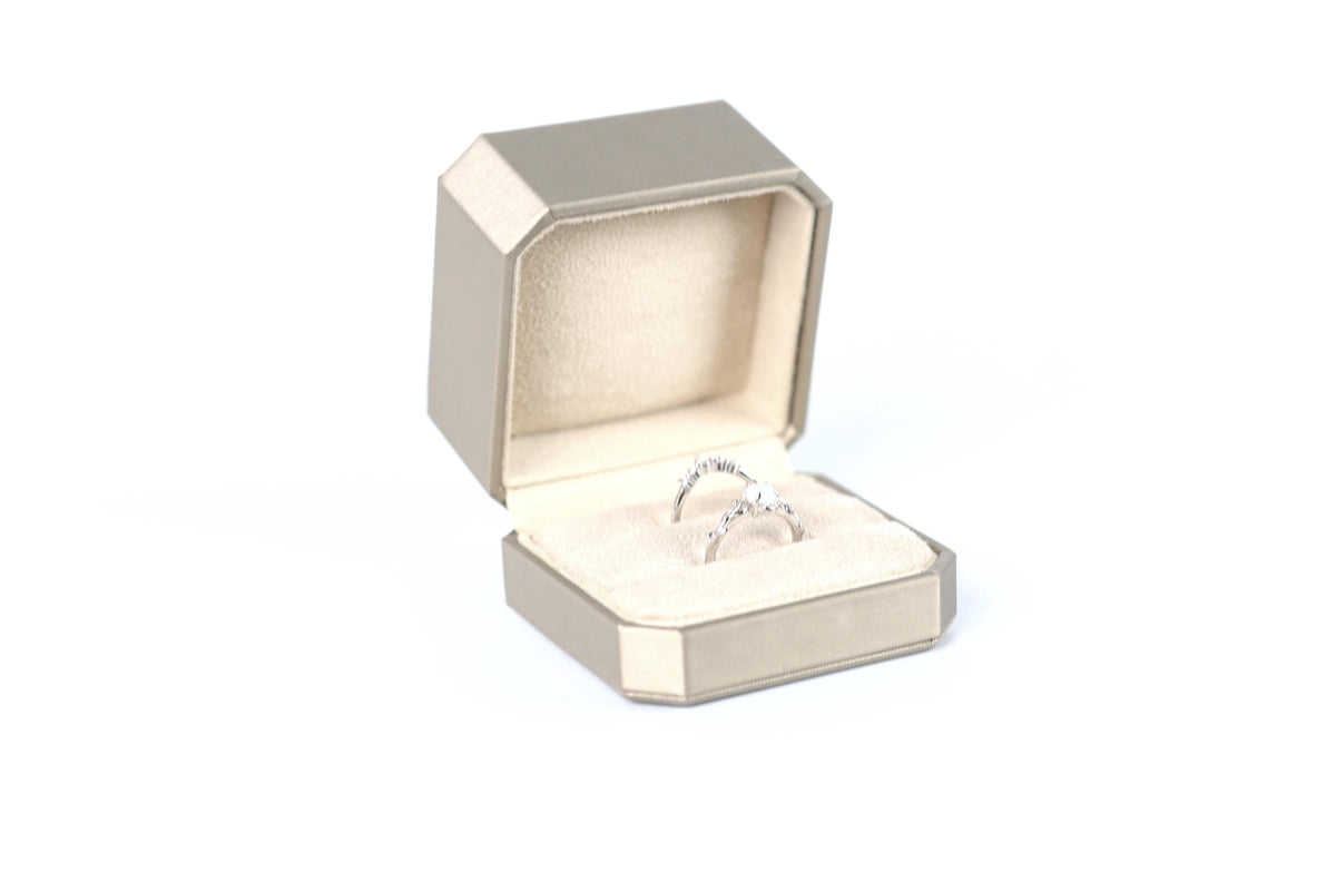 A silver ring is displayed in a gold jewelry box against a white background.