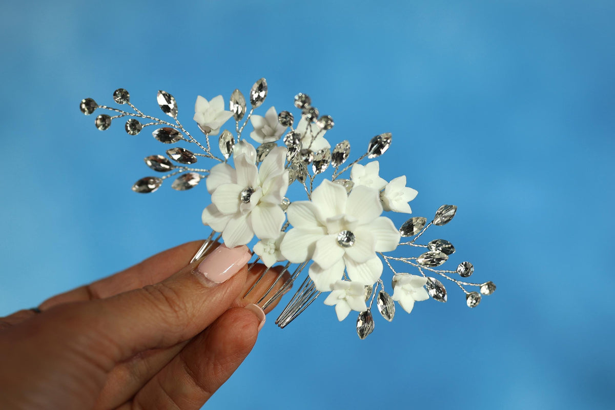A hand holding a decorative hair clip with white flowers and silver rhinestones against a blue background.