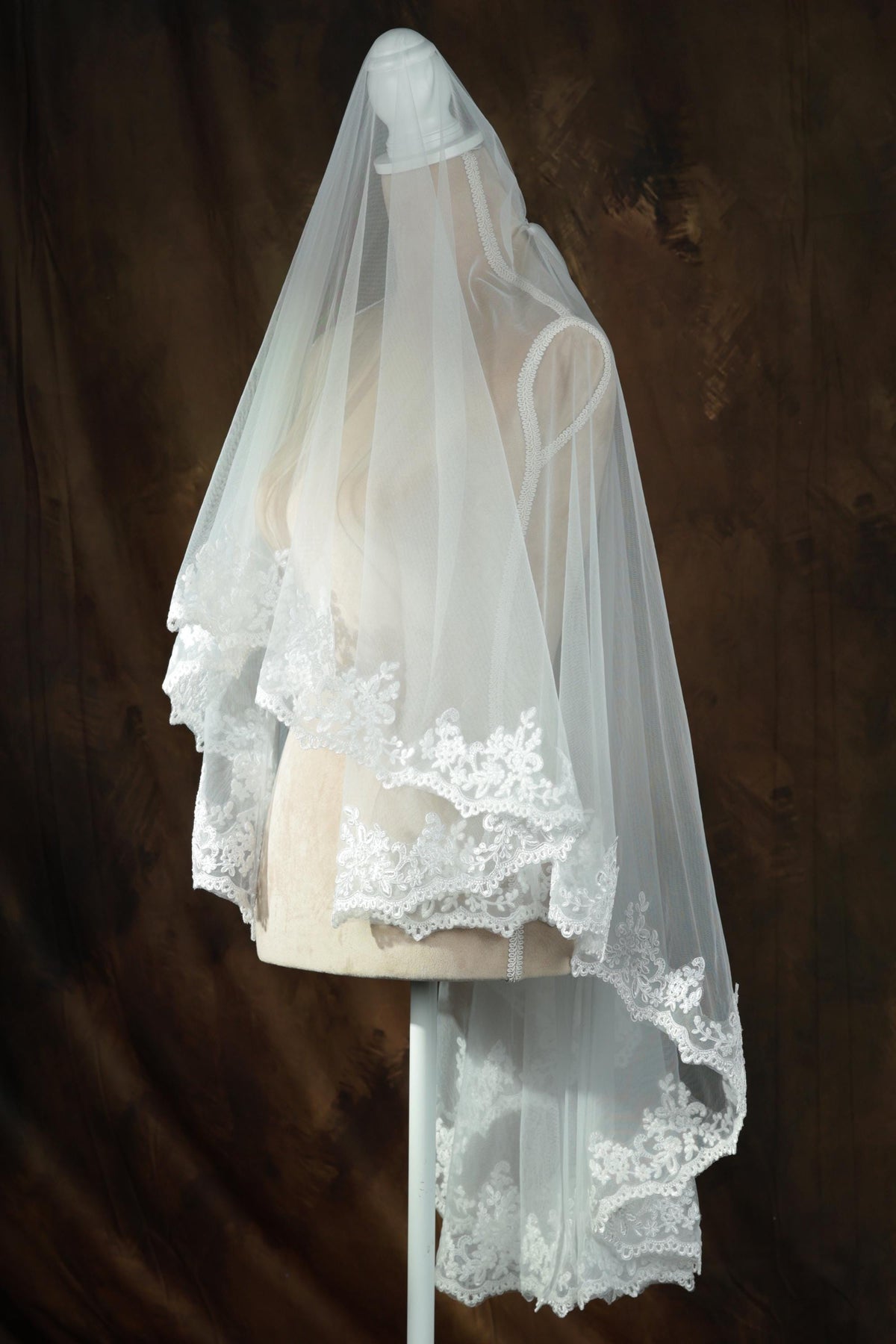 A white wedding veil with lace trim is displayed on a mannequin against a brown background.