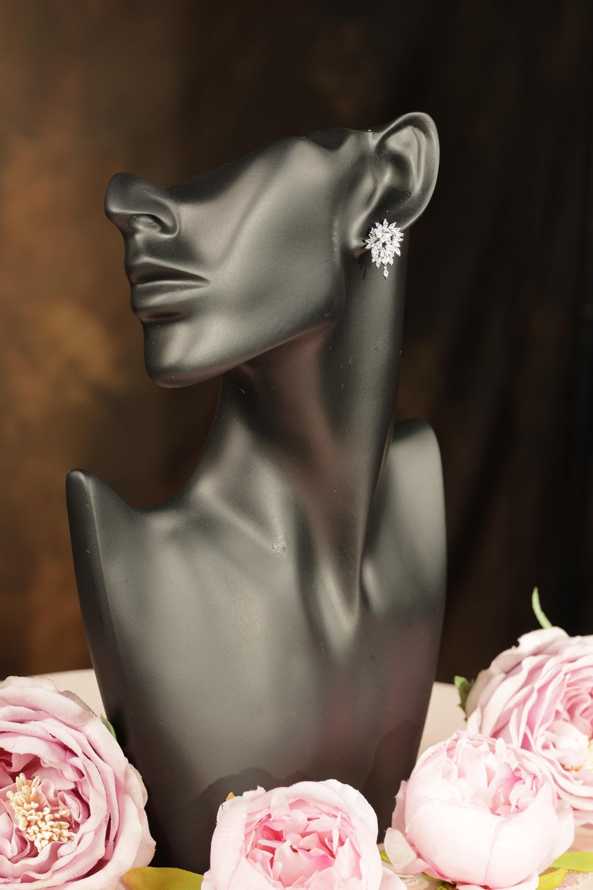A black bust form with a pair of earrings on it, placed next to a bouquet of pink roses.