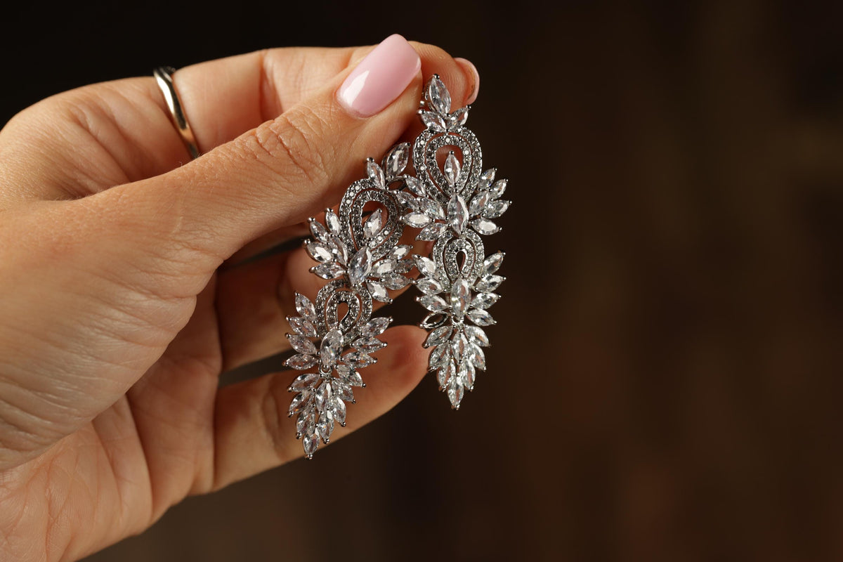 A hand holding a pair of elegant diamond earrings against a dark background.
