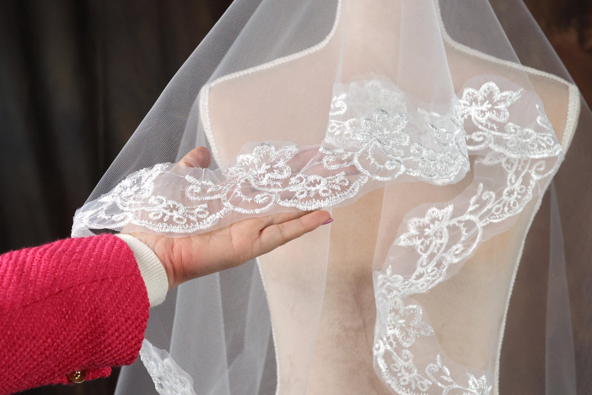 A hand is holding a white lace veil with intricate floral embroidery, which is displayed on a mannequin.