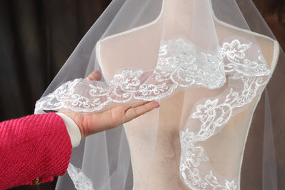 A hand is holding a white lace veil with intricate floral embroidery, which is displayed on a mannequin.