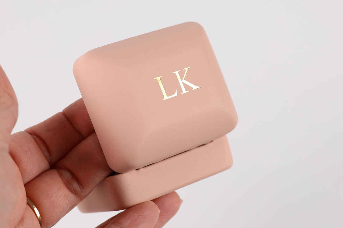 A hand holding a pink jewelry box with the initials "LK" engraved on it.