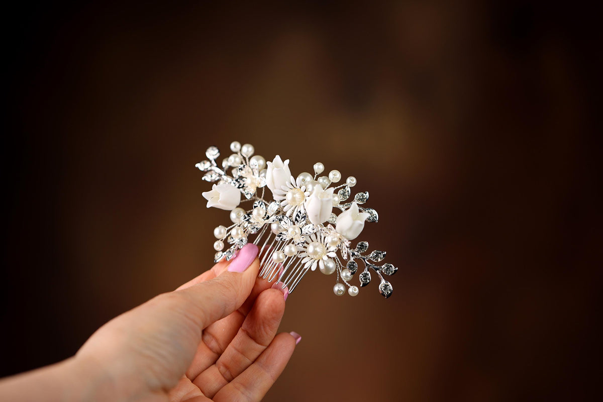 A hand holding a decorative hair clip with white flowers and pearls against a blurred brown background.