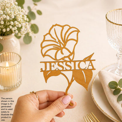 A hand holding a wooden cutout with the name 'JESSICA' on it, placed on a table with a candle, glass, and other decorative items.