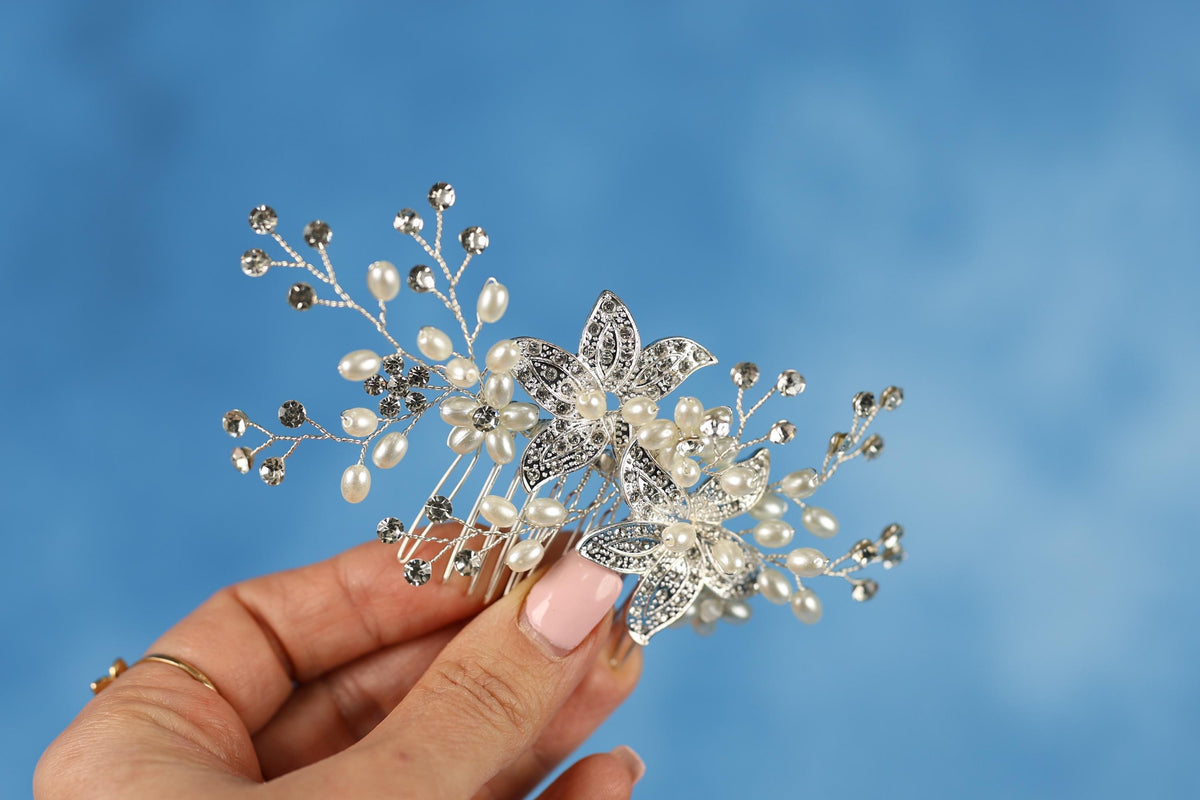 A hand holding a decorative hair accessory with a floral design and sparkling embellishments against a blue background.
