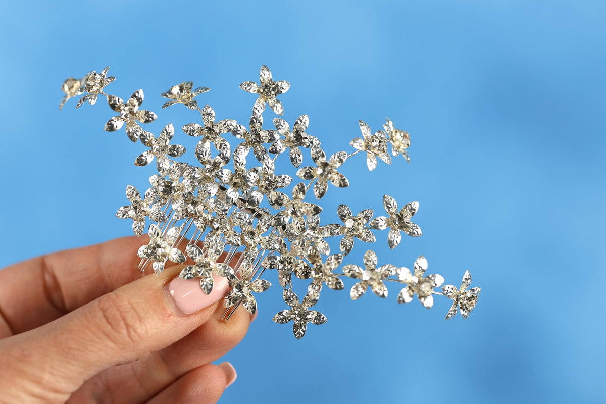 A hand holding a decorative silver flower-shaped hair accessory against a blue background.