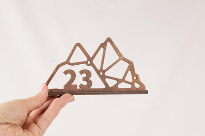 A hand is holding a wooden object that appears to be a mountain-shaped design with the number 23 on it.