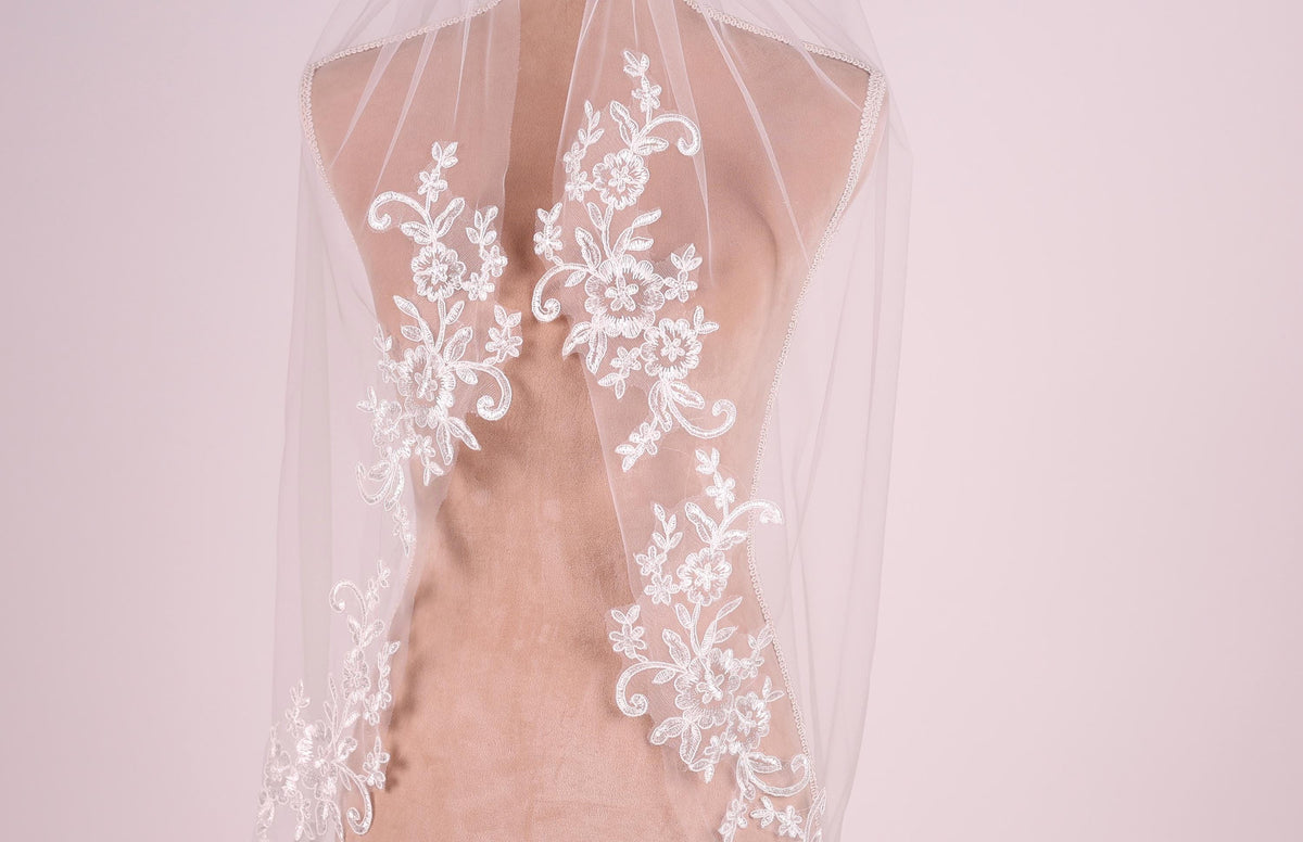 A close-up view of a delicate white lace veil with intricate floral embroidery, displayed on a mannequin or man-made figure.