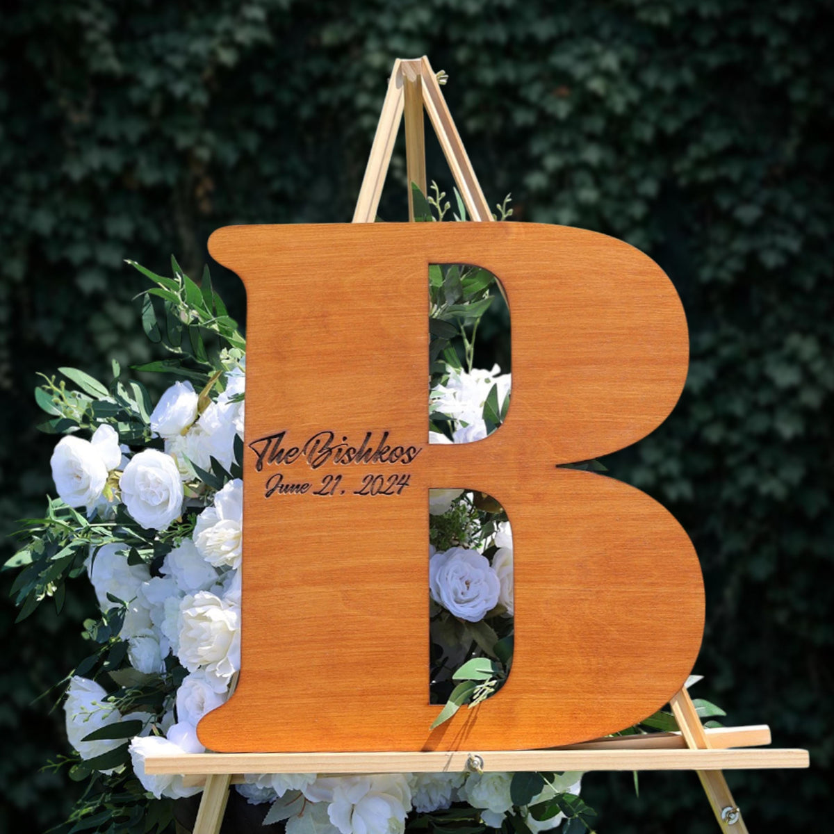a wooden letter 'B' with a floral arrangement of white roses and greenery behind it.