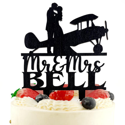A silhouette of a couple embracing an airplane on a cake topper, with the words "Mr & Mrs Bell" written below.