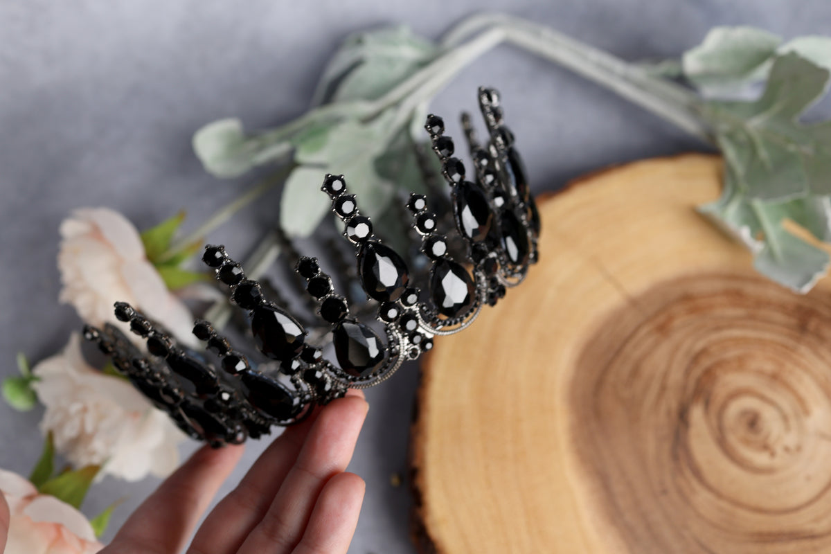 A hand is holding a black ornate tiara with black gemstones, surrounded by flowers and a wooden surface.